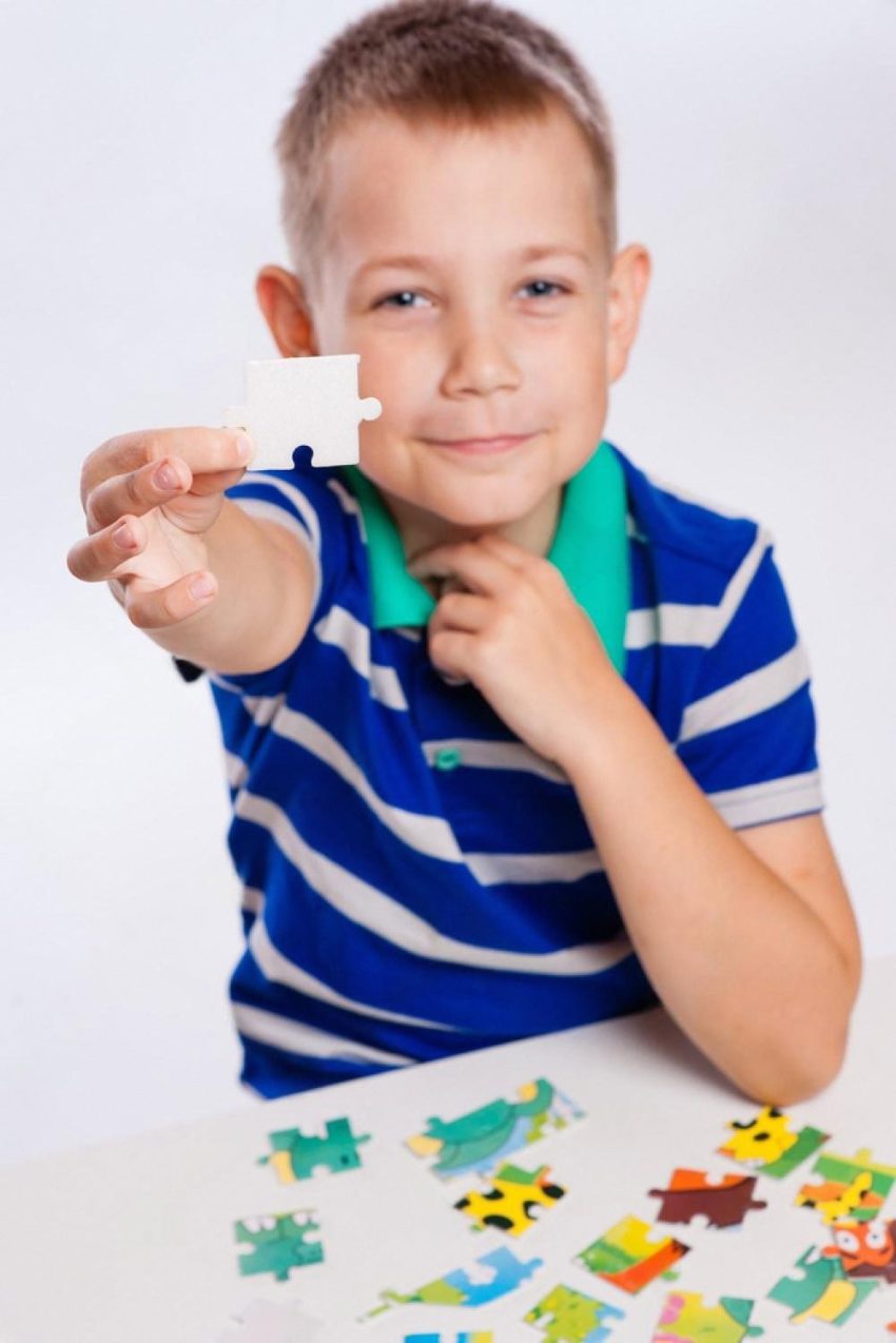 Young boy in a striped polo smiling and holding up a single white puzzle piece with colourful pieces on the table